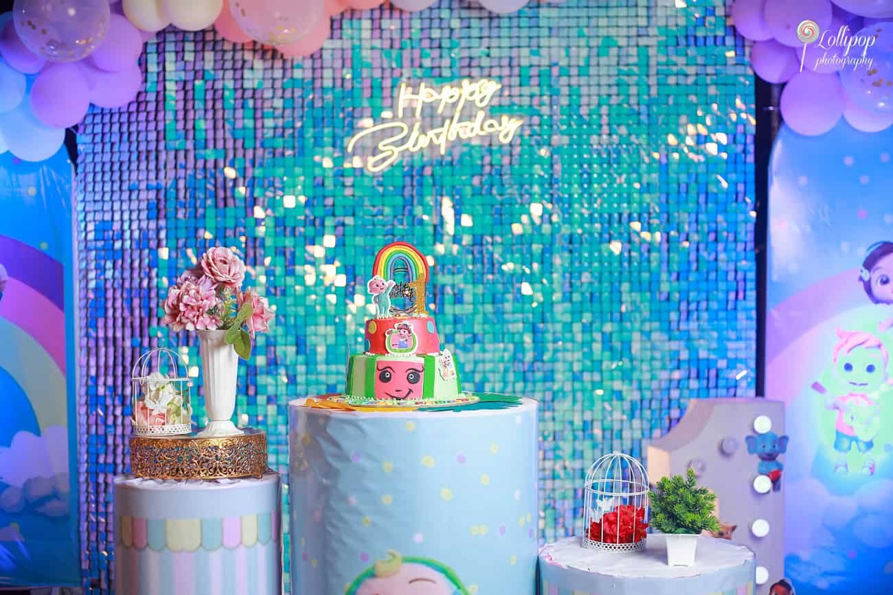 A beautifully crafted birthday cake on display under a canopy of blue and pink balloons with a shimmering blue backdrop reading "Happy Birthday" at a birthday event in Chennai.