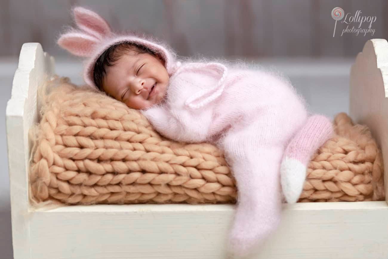 newborn-photoshoot-pink-bunny-outfit-chennai Joyful newborn in a pink bunny outfit, sleeping peacefully in a white cradle, photographed in Chennai by Lollipop Photography.