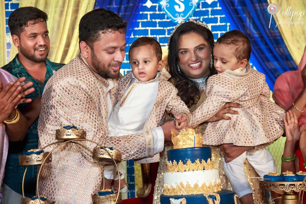 Smiling family moment with parents helping their twin boys cut a birthday cake at a grandly decorated blue and gold themed party in Chennai.
