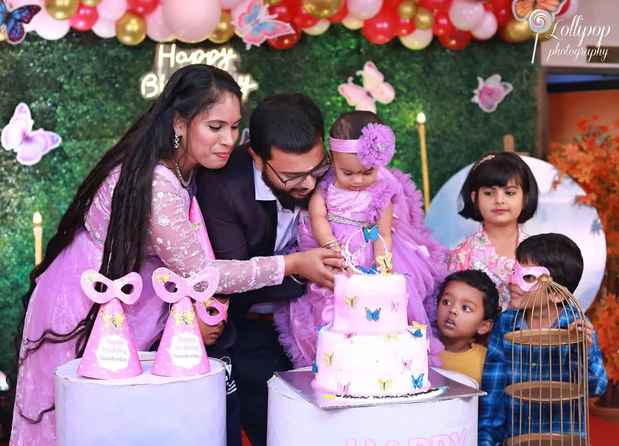 parents-helping-baby-girl-cut-birthday-cake-chennai Parents helping their baby girl cut her butterfly-themed birthday cake surrounded by family and friends. Birthday photoshoot by Lollipop Photography, Chennai.