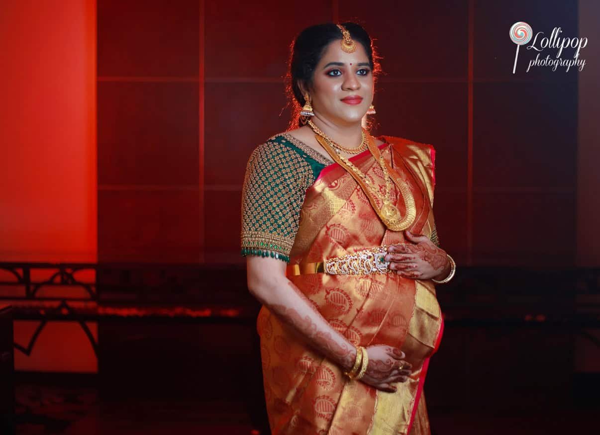 Hemalatha poses elegantly in an orange and green saree, adorned with gold jewelry, under warm lighting, showcasing the beauty of her baby shower celebration by Lollipop Photography.